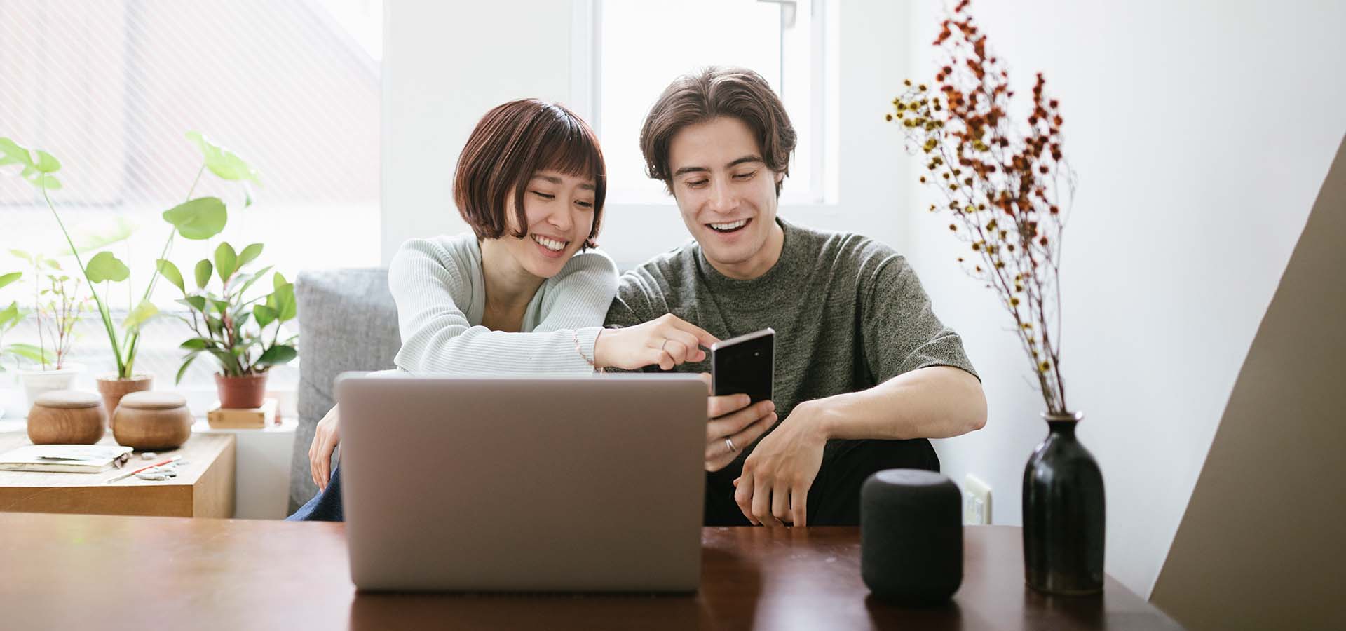 Two people next to an open laptop computer look at a mobile phone and smile.