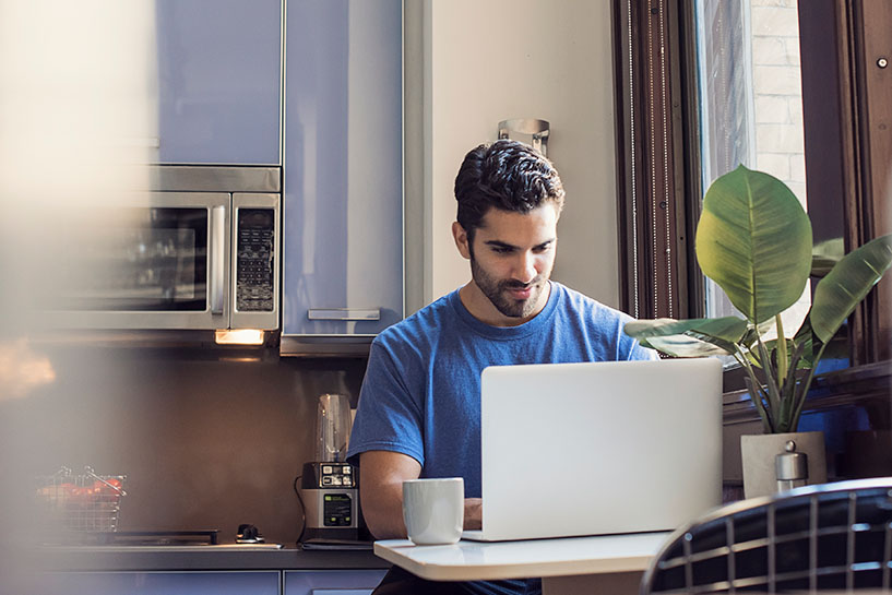 Un hombre sentado en su cocina y trabaja en una computadora portátil.