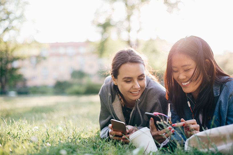 Two women lie in the grass and smile as they look at their mobile phones.