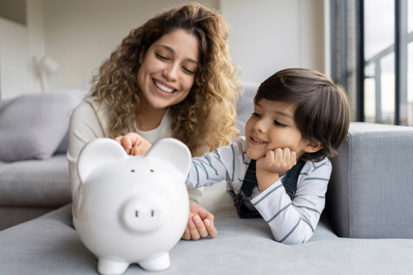 A woman watches as a young child puts a coin in a piggy bank.