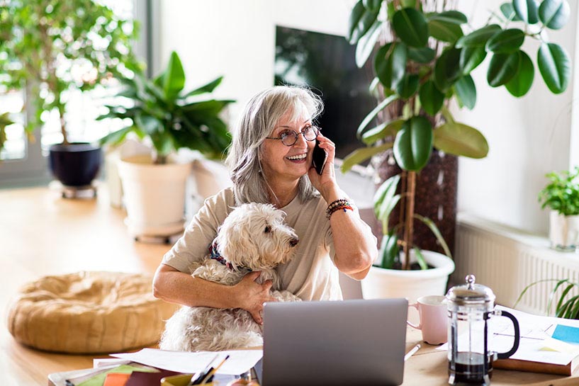 Una mujer y su perro sentados al lado de una laptop mientras la mujer habla en su teléfono móvil.