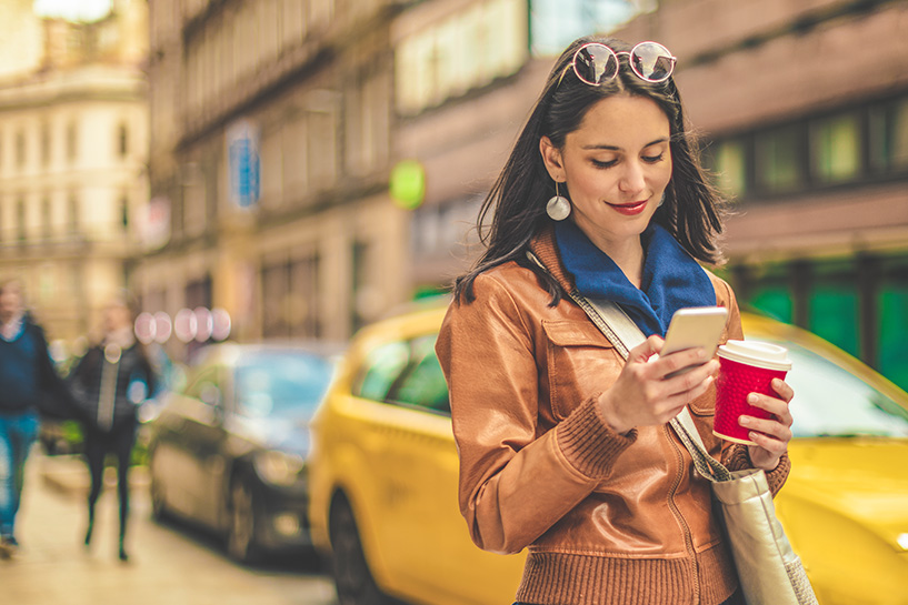 A woman holds a cup of coffee and uses her mobile phone.