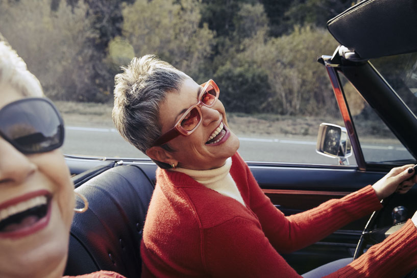 Two senior women smile and laugh as they drive in a convertible car.